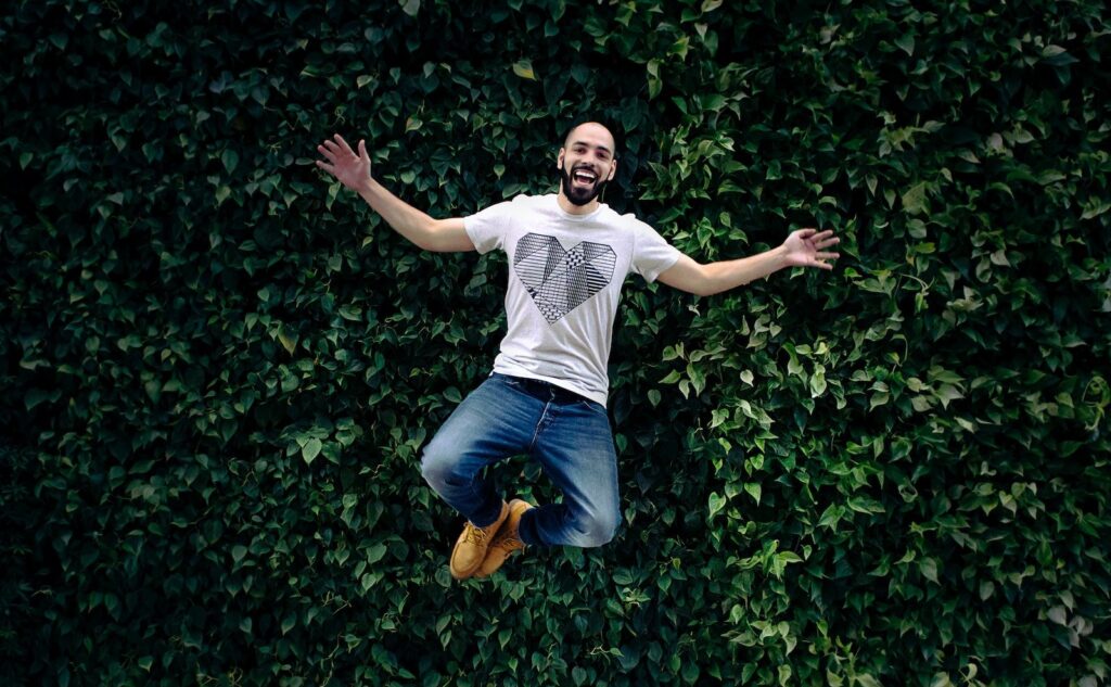 man wearing t-shirt and jeans jumpshot in front of a green hedge